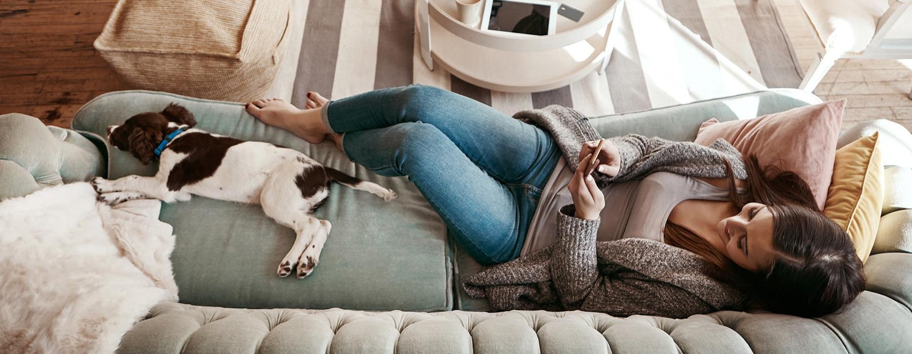 woman lies on a couch texting next to her dog